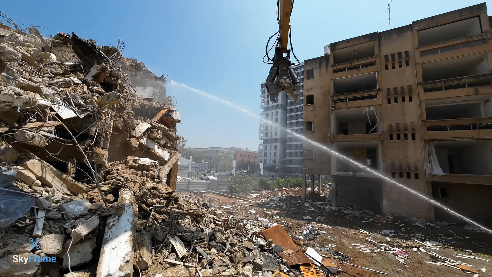 סילון מים מותז על ערימת הריסות ליד צבת ההריסה, ומאחור בניין ישן שעוד עומד ומגדל חדש