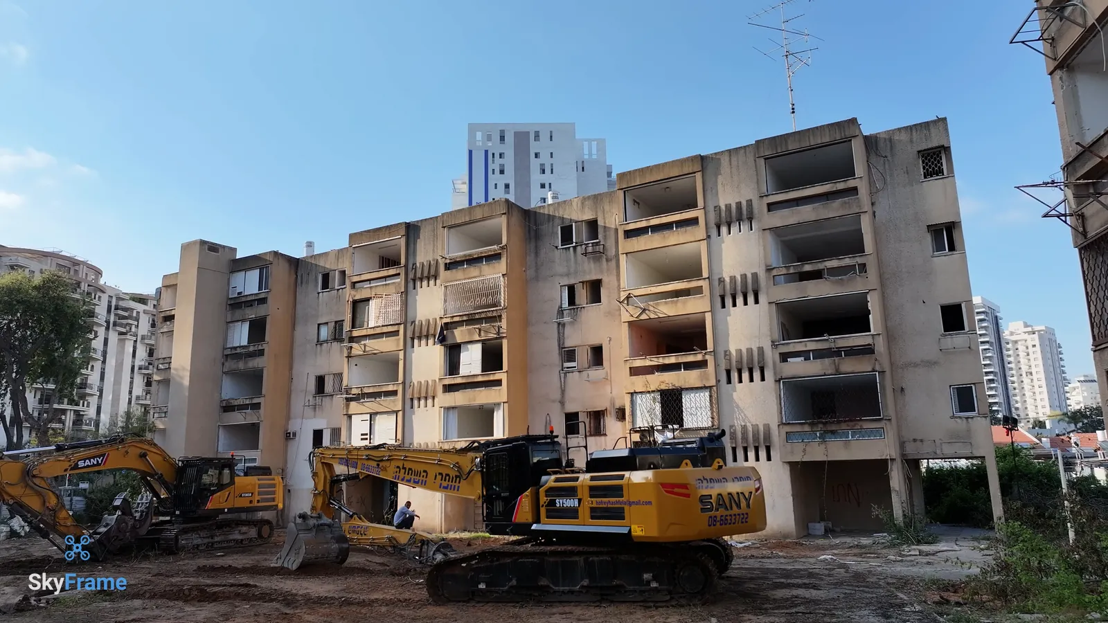 הבניין הישן בקריית אונו ביום טקס ההריסה, מחפרי ההריסה במגרש, ומגדלי מגורים חדשים ברקע