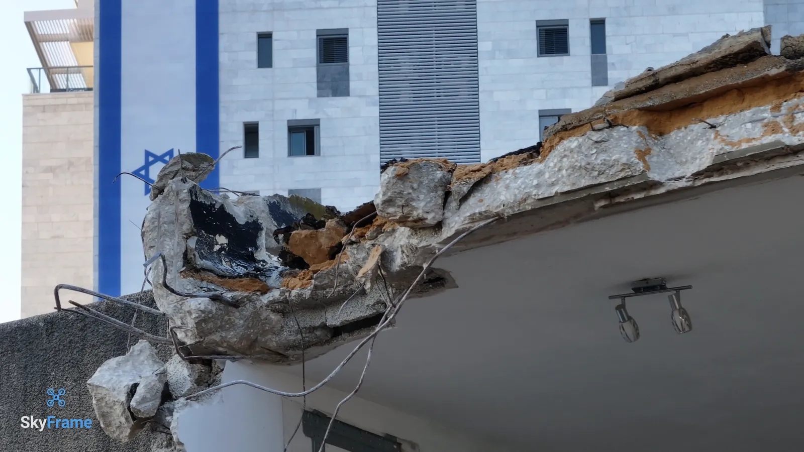 מרפסת בטון שבורה בבניין הישן, מנורה שעוד תלויה מתחתיה, וברקע מגדל מגורים חדש